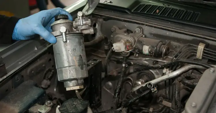 Ręka w niebieskiej rękawiczce wyjmuje z silnika metalowy element, co jest częścią wymiany filtra paliwa.