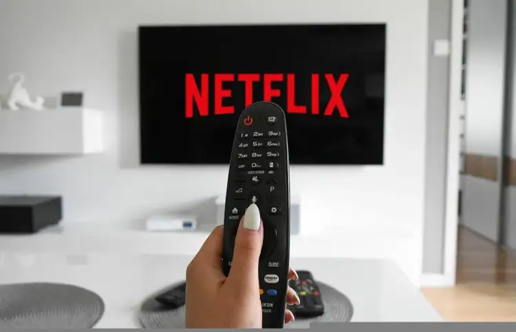 Dłoń trzymająca pilot do telewizora przed ekranem z logo Netflix.