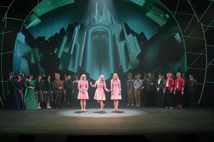 Trzy dziewczyny w różowych sukienkach na scenie, w tle zielony pałac Oz. To scena z musicalu "Wicked", który ma imponujący czas trwania.