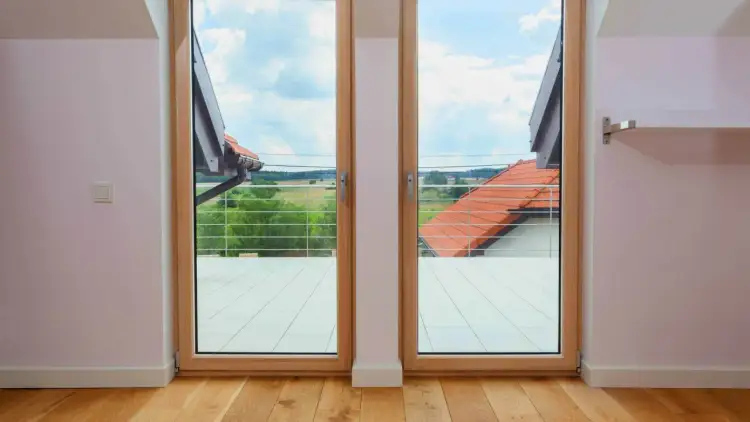 Drewniane okno balkonowe z widokiem na zielone pola i dachy. Zastanawiasz się, ile kosztuje okno balkonowe?