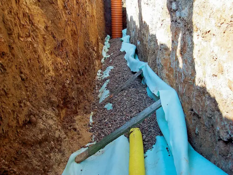 Wykopy pod drenaż wokół starego domu. Widać rury drenarskie, żwir i geowłókninę.