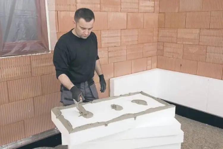 Pracownik nakłada zaprawę na styropian, przygotowując go do montażu. To kluczowy etap, jak przymocować belkę drewnianą do ściany ocieplonej styropianem.