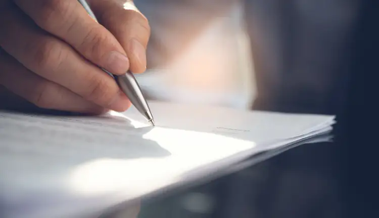 Dłoń z długopisem nad stosem dokumentów, być może odpis wyroku sądu online.