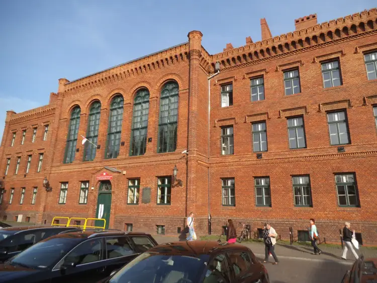 Ceglana kamienica z zielonymi oknami, gdzie w wieku 19 lat kończy się liceum. Ludzie idą chodnikiem.