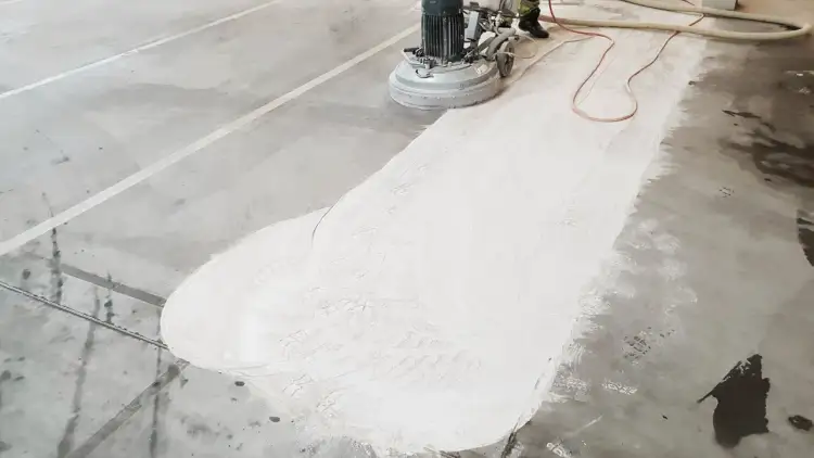 Maszyna szlifuje betonową posadzkę, tworząc biały pył. To przygotowanie do wypełnienia ubytków w posadzce betonowej.