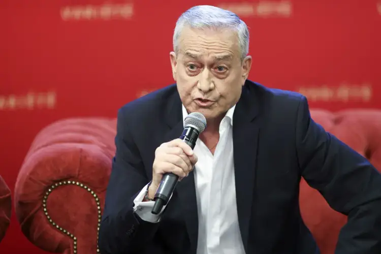 Janusz Szydłowski przemawia do mikrofonu, siedząc na czerwonej, pikowanej kanapie.