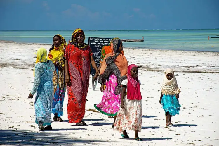 Kobiety i dziewczynki w kolorowych strojach spacerują po piaszczystej plaży na Zanzibarze. W tle turkusowe morze i błękitne niebo.