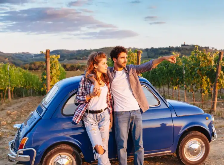 Para młoda przy niebieskim Fiat 500, planując podróż po Włoszech samochodem, wskazuje kierunek na tle winnic.