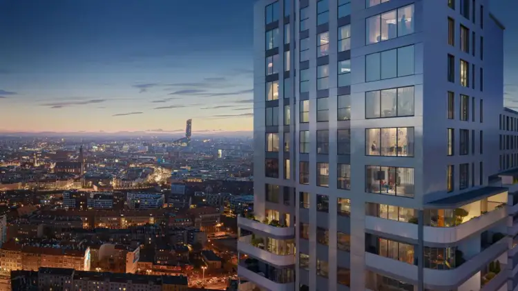 Nowoczesne osiedle apartamentowców w mieście nocą. Widać oświetlone okna, balkony z zielenią i panoramę miasta w tle.