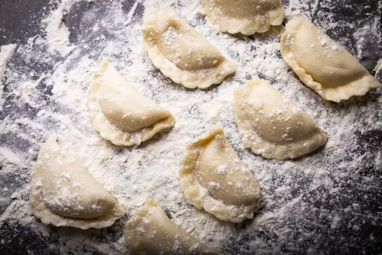 Surowe pierogi leżą na blacie oprószonym mąką. Jaki typ mąki na pierogi? Ta wygląda na pszenną, idealną do domowych wypieków.