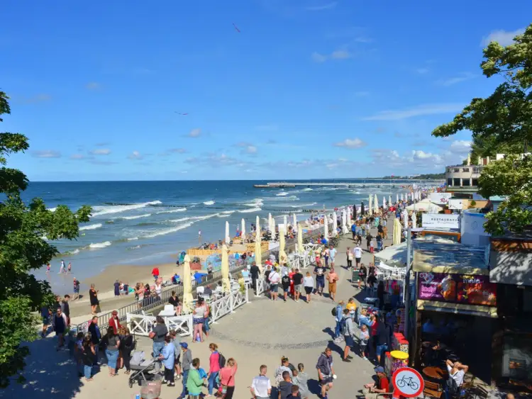 Tłum ludzi spaceruje promenadą w Kołobrzegu, obok plaży i restauracji. Zastanawiasz się, ile kosztuje klimatyczne w Kołobrzegu?