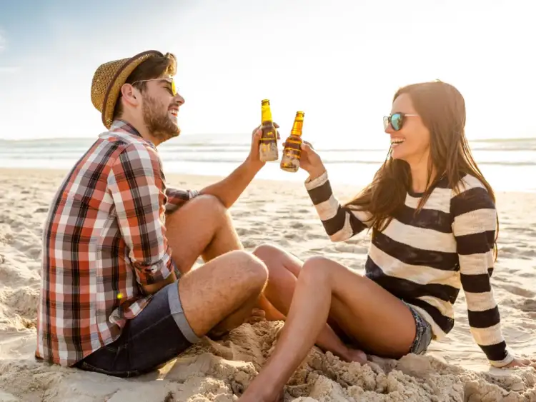 Para dwojga świętuje romantyczny weekend dla dwojga nad morzem, wznosząc tosty piwem na plaży.