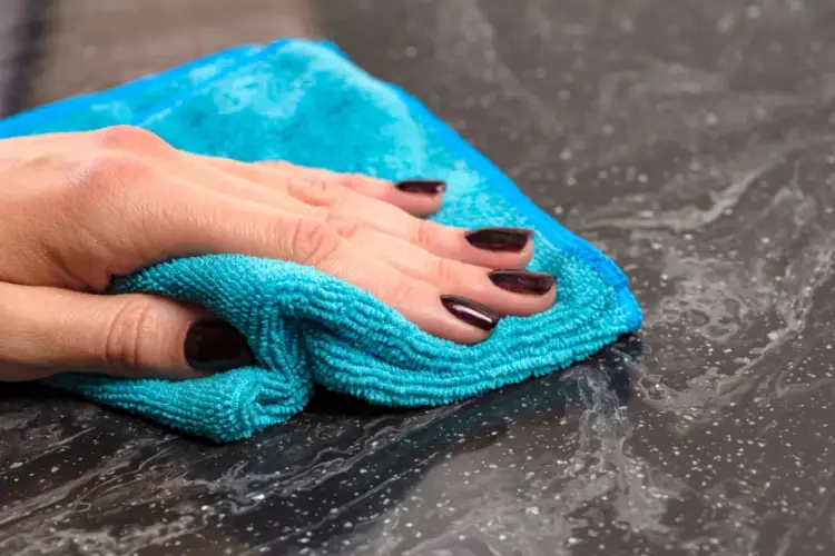 Dłoń z ciemnymi paznokciami czyści ciemny blat turkusową ściereczką. Dowiedz się, jak usunąć plamy z farby do włosów z mebli.
