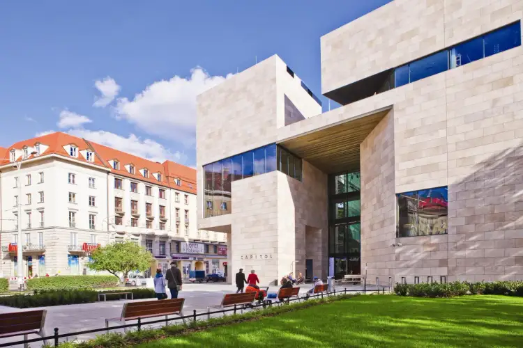 Nowoczesny budynek Capitol we Wrocławiu, z napisem "CAPITOL" na fasadzie, otoczony zielenią i ławkami.