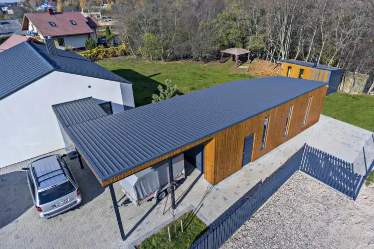 Nowoczesny garaż z drewnianą elewacją i dachem z blachy trapezowej. Obok stoi przyczepa.