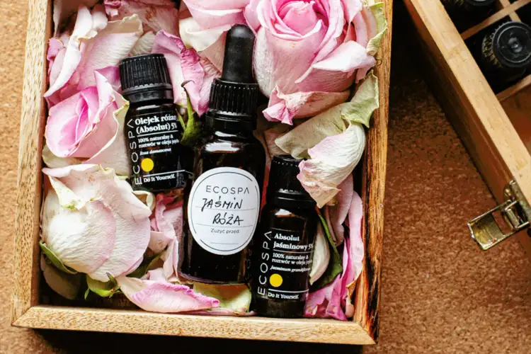 Naturalne olejki różany i jaśminowy w drewnianej skrzynce wśród płatków róż. Dowiedz się, jak oszczędzić na perfumach, wybierając esencje.