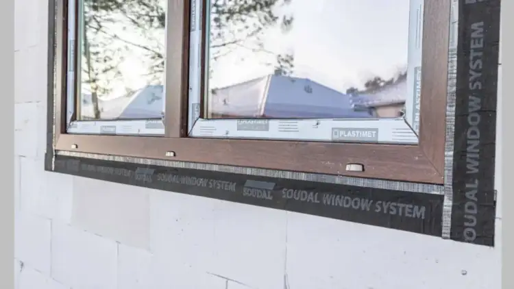 Ciepły montaż po zamontowaniu okien. Widoczna taśma Soudal Window System wokół drewnianej ramy okiennej w białej ścianie z bloczków.