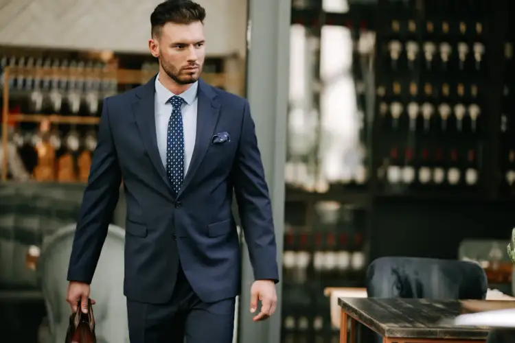 Elegancki mężczyzna w granatowym garniturze, idealny przykład, jak się ubrać do teatru na komedię. W dłoni trzyma brązową teczkę.