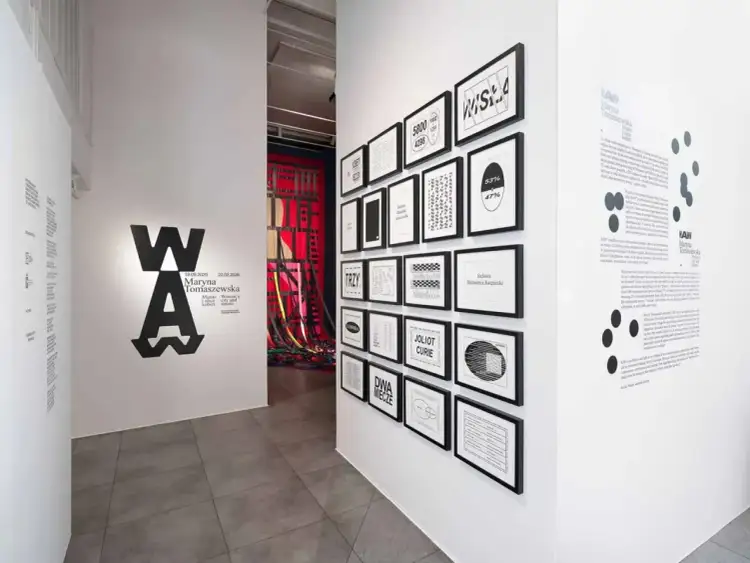 Wystawa w muzeum sztuki nowoczesnej prezentuje prace Maryny Tomaszewskiej. Na ścianie widnieje logo WA oraz grafiki z datami i tytułami.