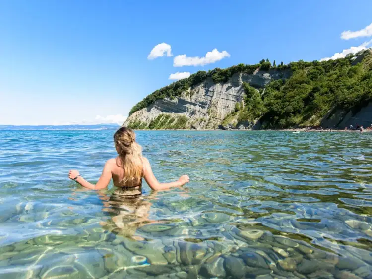Kobieta w wodzie, na tle klifu i błękitnego nieba. Widok potwierdza, że Słowenia ma dostęp do morza.