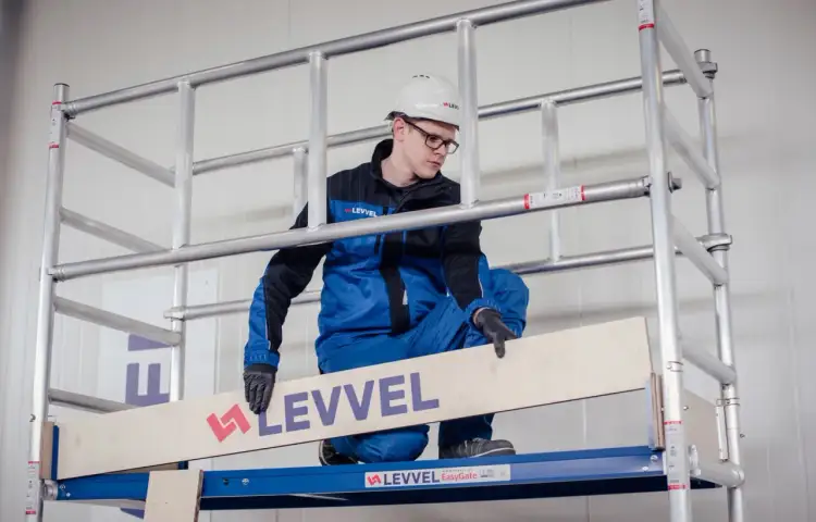 Mężczyzna w kasku i niebieskim kombinezonie demonstruje, jak złożyć rusztowanie aluminiowe, umieszczając drewnianą deskę.