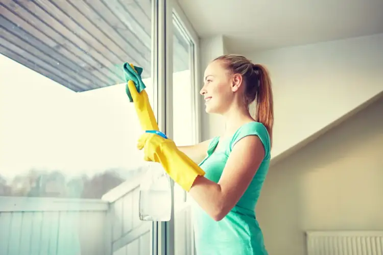 Uśmiechnięta dziewczyna w żółtych rękawiczkach myje okno, używając domowego sposobu: mycie okien octem.