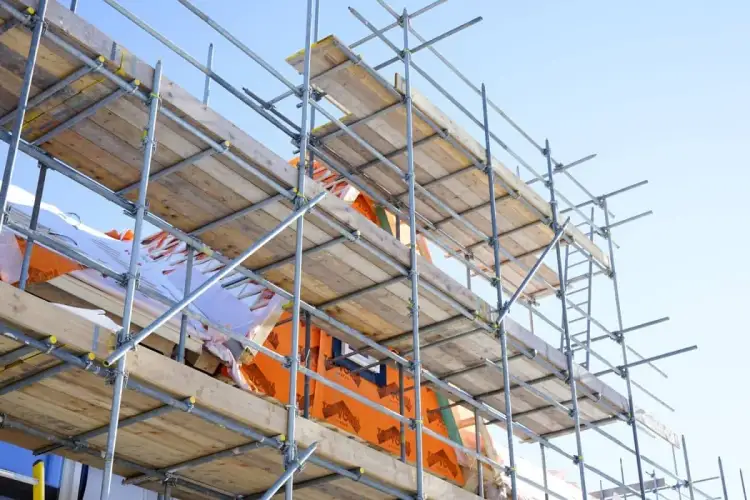 Konstrukcja rusztowania z drewna i metalu, gotowa do pracy. Widać deski i pomarańczową membranę.