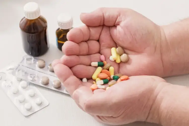 Dłonie trzymające garść kolorowych tabletek i kapsułek, obok buteleczki z lekami i blistry.