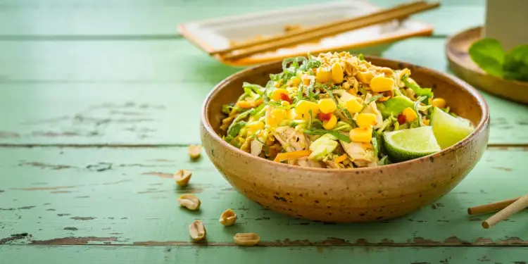 Kolorowa sałatka tajska z kurczakiem, kukurydzą, orzeszkami ziemnymi i limonką w glinianej misce na zielonym, drewnianym stole.