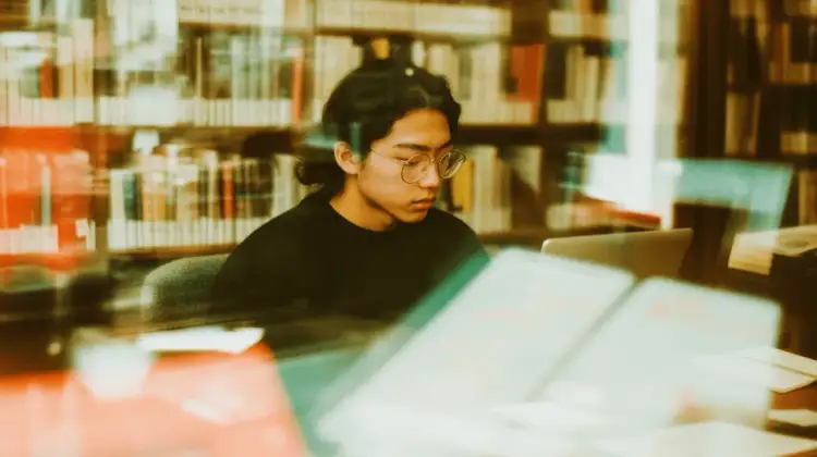 Student w okularach pochylony nad laptopem w bibliotece, przygotowuje podanie o indywidualny tok studiów uzasadnienie przykład.