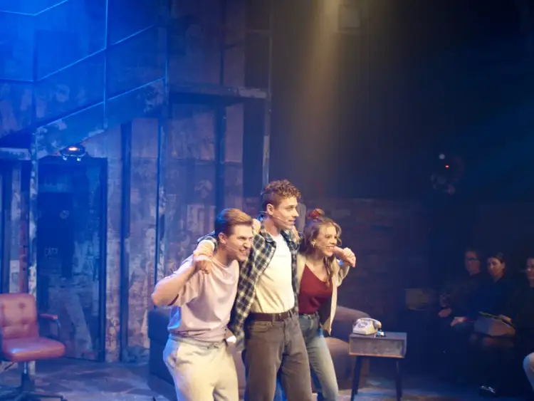 Trzech młodych ludzi na scenie, w tle ceglana ściana i niebieskie światła. Może to scena z musicalu "Tik Tik Bum" w Rzymie?