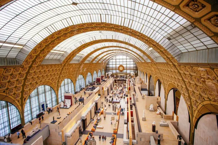 Wnętrze muzeum d'Orsay z imponującym szklanym sufitem i łukami. Zwiedzający spacerują wśród rzeźb i dzieł sztuki.
