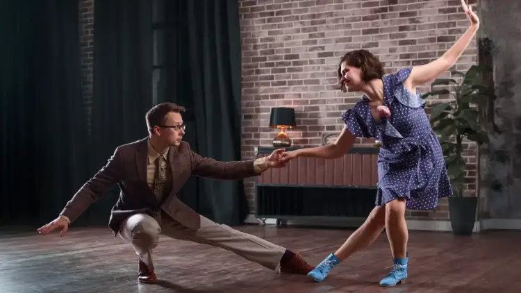 Para, ćwicząca naukę tańca towarzyskiego, w eleganckim brązowym garniturze, trzyma dłoń partnerki w granatowej sukience w kropki.