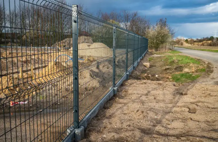 Trwa montaż ogrodzenia panelowego. Nowy płot z betonową podmurówką oddziela plac budowy od drogi i terenów zielonych.