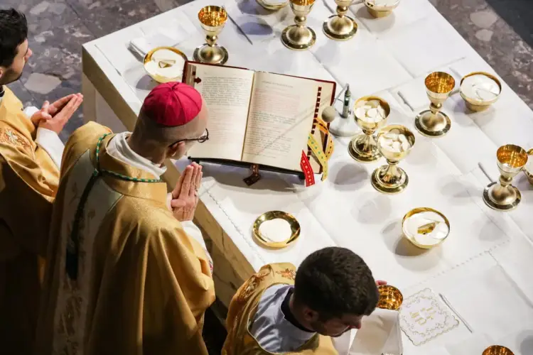 Biskup w czerwonej piusce i złotej szacie modli się nad otwartą księgą podczas drugiej modlitwy eucharystycznej. Wokół kielichy z hostiami.