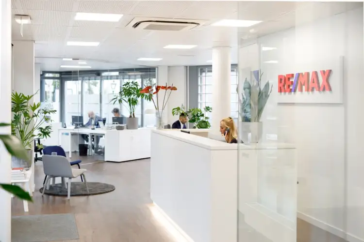 Nowoczesne biuro nieruchomości RE/MAX. Pracownicy obsługują klientów przy recepcji i biurkach.