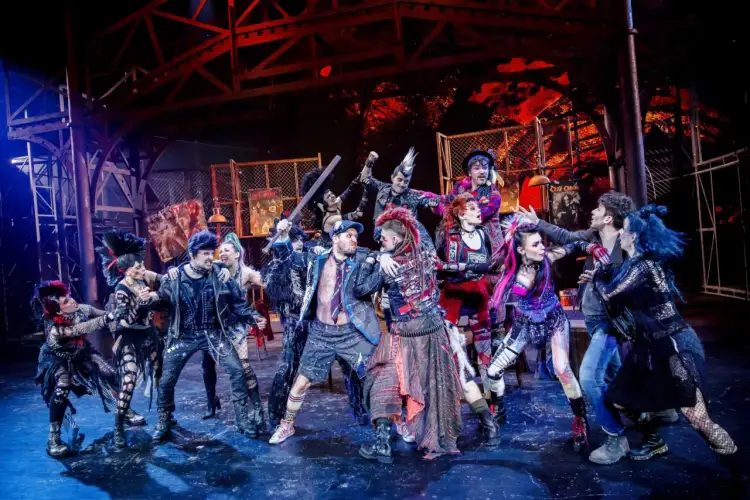 Grupa punkowych postaci na scenie, w tym z irokezami i w skórzanych kurtkach, w dynamicznej scenie z musicalu "We Will Rock You".