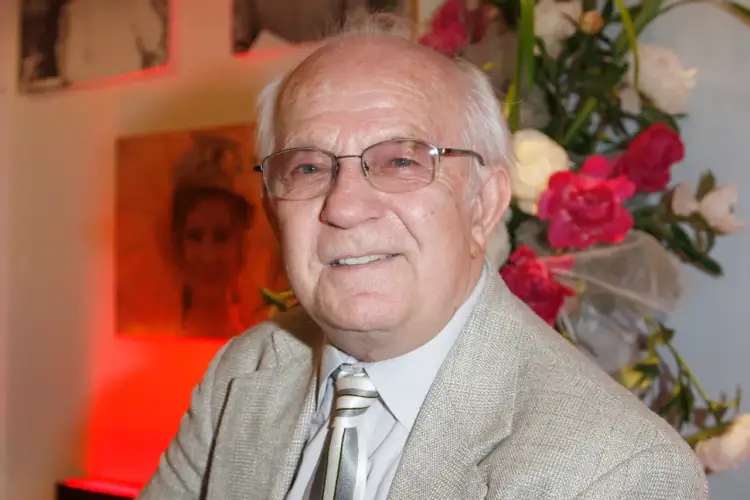 Uśmiechnięty Jerzy Antczak, reżyser znany z niezapomnianych filmów, w eleganckim garniturze, na tle bukietu kwiatów i portretu.