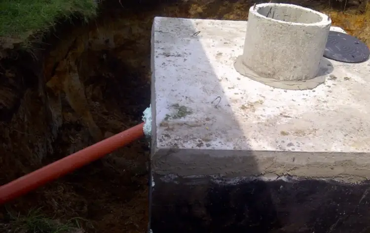 Pomarańczowa rura do szamba wychodzi z wykopu i wchodzi w betonową konstrukcję. Obok betonowy krąg studzienki.