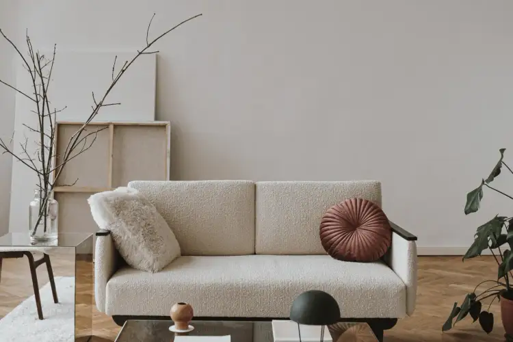 Przytulna aranżacja małego mieszkania: kremowa kanapa z ozdobnymi poduszkami, szklany stolik z gałązkami i rośliną, tworzą stylową przestrzeń.