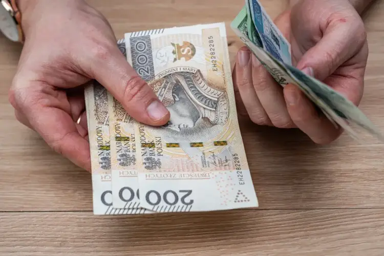 Dłonie liczą polskie banknoty. Czy mieszkaniówka wlicza się do dochodu? Pieniądze na drewnianym blacie.