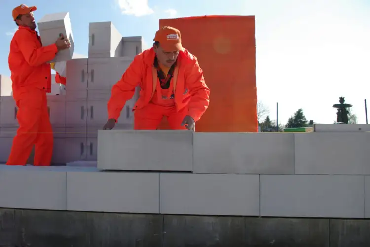 Dwóch budowlańców w pomarańczowych strojach układa białe pustaki. Jeden niesie pustak, drugi go kładzie.