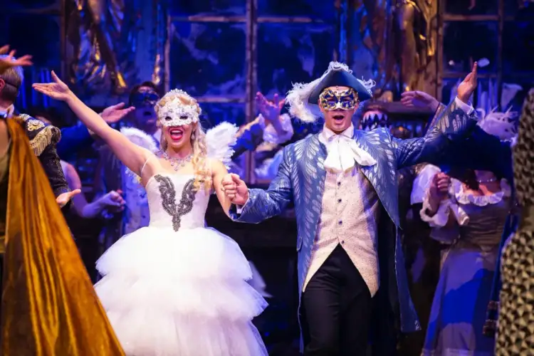 Upiór w Operze Warszawa: para w maskach i strojach z epoki tańczy na balu. Ona w białej sukni i z anielskimi skrzydłami, on w błękitnym fraku.