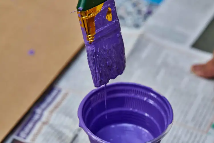 Pędzel zanurzony w fioletowej farbie, z której ściekają krople do plastikowego pojemnika.
