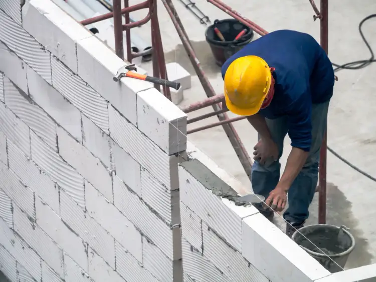 Budowlaniec układa bloczki z betonu komórkowego, dbając o precyzyjne wymiary ściany. Młotek i kielnia czekają na użycie.
