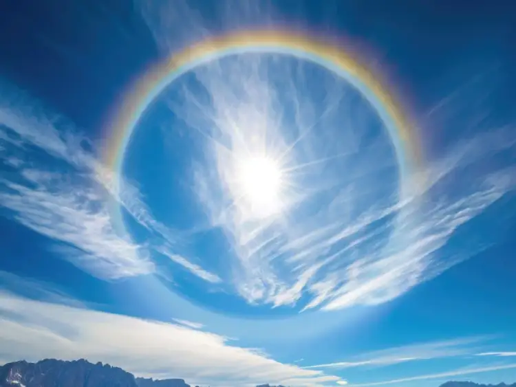 Słońce otoczone tęczowym pierścieniem, przypominającym efekt halo przy astygmatyzmie. Na niebie delikatne chmury i zarys gór.
