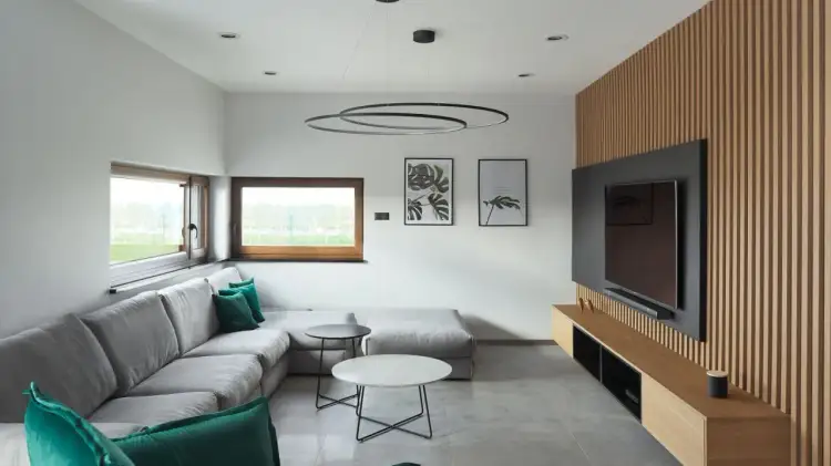 Nowoczesny salon z szarą kanapą i zielonymi poduszkami. Dekor na ścianie w salonie to drewniana boazeria i telewizor.