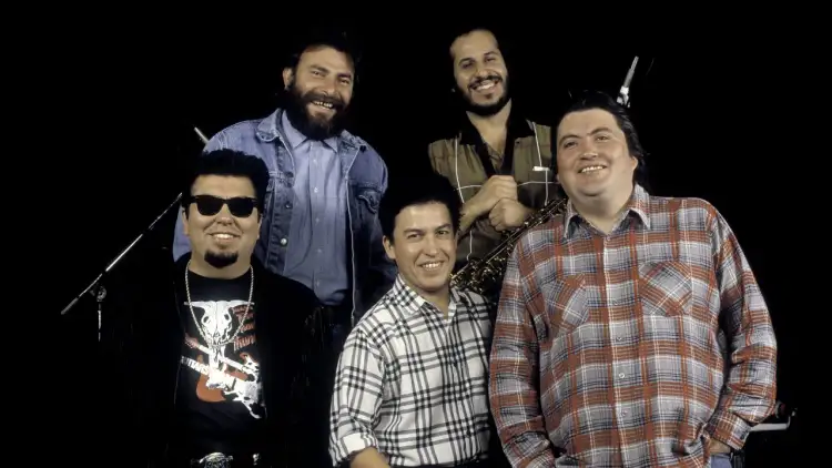 Los Lobos, native sons, pose together. One man wears sunglasses and a fringed jacket, another a denim jacket, and two sport plaid shirts. A saxophone is visible.