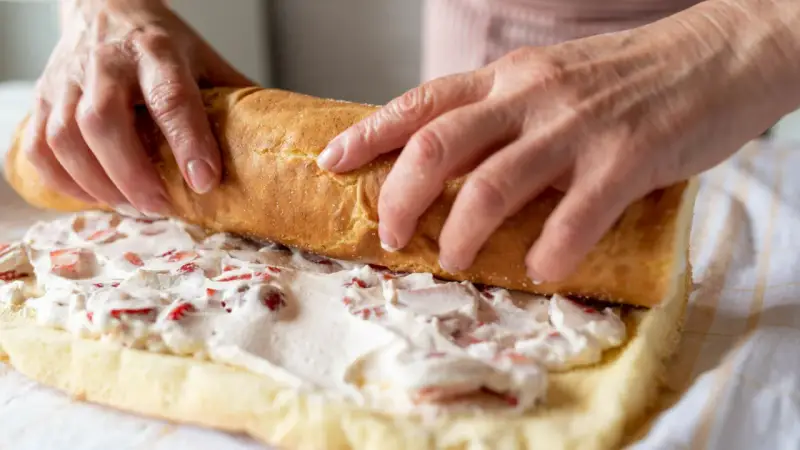 Ręce zwijają biszkopt z kremem i truskawkami. To najprostsza rolada biszkoptowa, która zachwyci smakiem.