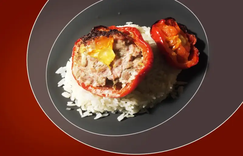 Poivrons rouges farcis à la viande, servis sur un lit de riz blanc. Une sauce légère accompagne ce plat, rappelant les saveurs des tomates farcies.